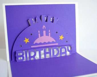 May include: A purple pop-up greeting card featuring the text HAPPY BIRTHDAY in cut-out letters. The design includes a light purple cake with three lit candles, two yellow stars, and small circular cut-outs on a semi-circular purple background.