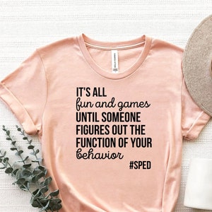 May include: A peach-colored t-shirt with the text "IT'S ALL fun and games UNTIL SOMEONE FIGURES OUT THE FUNCTION OF YOUR behavior #SPED" printed in black.