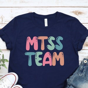 May include: A navy blue t-shirt with the text "MTSS TEAM" in rainbow colors. The shirt is laid flat on a white wooden surface with a pair of white sneakers and blue jeans.