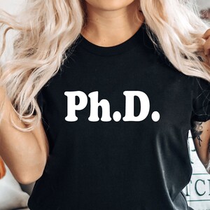May include: Black t-shirt with the white text "Ph.D." printed on the front. The shirt is a crew neck style. A decorative pillow with a pumpkin design is visible in the background.
