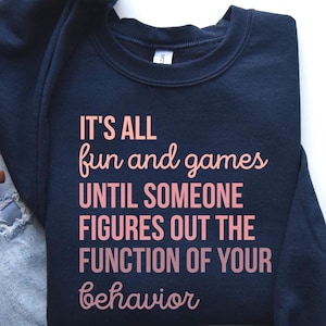 May include: A navy blue sweatshirt with the text "It's all fun and games until someone figures out the function of your behavior" printed in pink.