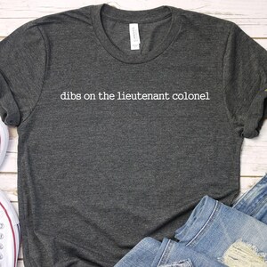 May include: Dark gray t-shirt with the text "dibs on the lieutenant colonel" in white. The shirt is paired with white sneakers and blue jeans on a white wooden surface. A sprig of greenery is also present.