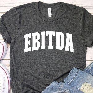 May include: A dark gray t-shirt with the word "EBITDA" printed in white on the front. The shirt is laid flat on a white wooden surface with a pair of white sneakers and blue jeans.
