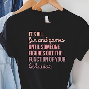 May include: Black t-shirt with a pink graphic that reads "It's all fun and games until someone figures out the function of your behavior."
