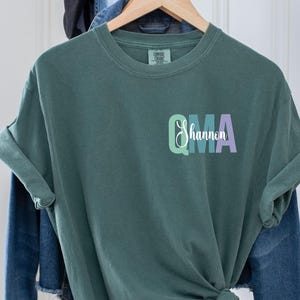 May include: A dark green t-shirt with a colorful "QMA" graphic and the text "Shannon" underneath. The shirt is folded and hanging on a hanger.