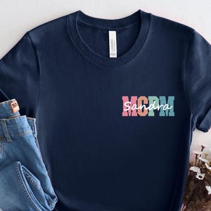 May include: A navy blue t-shirt with the word "MOM" in colorful block letters and the name "Sandra" in a script font. The shirt is paired with a pair of blue jeans. A bouquet of white flowers is placed to the side.