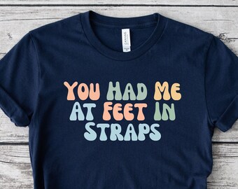 May include: Navy blue t-shirt with the text "YOU HAD ME AT FEET IN STRAPS" in retro-style lettering. The text is in orange, peach, green, and light blue. The shirt is knotted at the bottom.