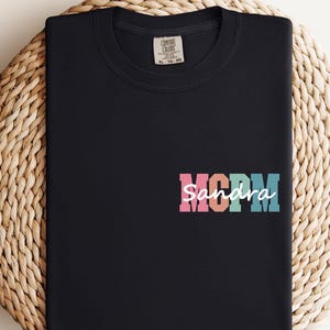 May include: Black t-shirt with the word "MOM" in pink, orange, teal, and blue, with the name "Sandra" in white script below. The shirt is folded on a woven, circular surface.