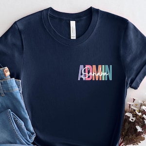 May include: A navy blue t-shirt with the word "ADMIN" in colorful text on the front. The shirt is paired with a pair of blue jeans. A small bouquet of white flowers is placed next to the shirt and jeans.