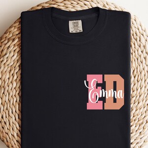 May include: Black t-shirt with a personalized design. The design features the initials "ED" in pink and orange, with the name "Emma" in white script. The shirt is folded on a woven, circular placemat.