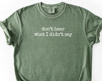 May include: Olive green t-shirt with rolled sleeves. The shirt features the text "don't hear what I didn't say" in white, centered on the front. The shirt is laid flat on a white surface.