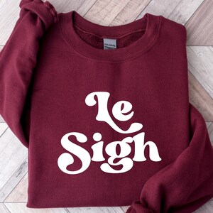 May include: A maroon sweatshirt with the text "Le Sigh" in white retro-style lettering.