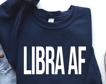May include: A navy blue sweatshirt with the text "LIBRA AF" printed in white on the front. The sweatshirt is laid flat on a white surface with a pair of light blue jeans and a brown coffee mug.