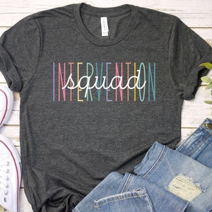 May include: A dark gray t-shirt with the text "Intervention Squad" printed in rainbow colors.