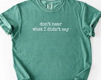 May include: A teal-colored t-shirt with the phrase "don't hear what I didn't say" printed in white. The shirt is folded and has rolled-up sleeves, showcasing a casual style. The fabric appears soft and comfortable.