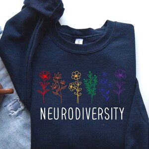 May include: A navy blue sweatshirt with a rainbow floral design and the text "Neurodiversity". The sweatshirt is laid flat on a white surface with a pair of grey sneakers and blue jeans.