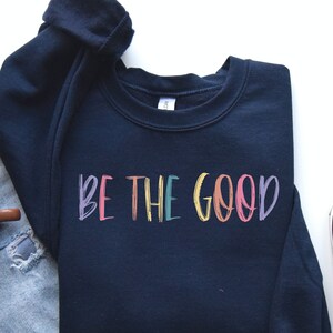 May include: A navy blue sweatshirt with the text "BE THE GOOD" in rainbow colors. The sweatshirt is laid out on a white surface with a pair of light blue jeans and a brown coffee mug.