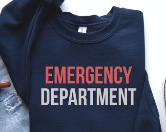 May include: A navy blue sweatshirt with the text "EMERGENCY DEPARTMENT" in red and white lettering. The sweatshirt is laid out on a white surface with a pair of light gray sneakers and a pair of blue jeans.