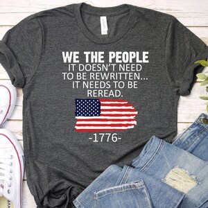May include: A dark gray t-shirt with the text "WE THE PEOPLE IT DOESN'T NEED TO BE REWRITTEN... IT NEEDS TO BE REREAD. -1776-" and a distressed American flag graphic.