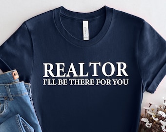 May include: A navy blue t-shirt with white text that reads "REALTOR I'LL BE THERE FOR YOU". The shirt is folded and displayed on a white background with a pair of blue jeans and white flowers.