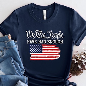 May include: A navy blue short-sleeve t-shirt featuring the white text We The People HAVE HAD ENOUGH above a graphic of the American flag with a distressed, brushstroke-style finish. The shirt is paired with blue denim jeans with distressed details.