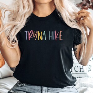 May include: A black t-shirt with the text "TRYNA HIKE" in rainbow colors. The text is written in a casual, handwritten style.