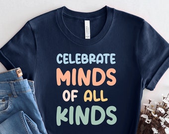 May include: A navy blue t-shirt with the text "Celebrate Minds of All Kinds" in colorful letters. The shirt is folded and displayed on a white background with a pair of blue jeans and a bouquet of white flowers.