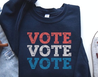 May include: Navy blue sweatshirt with the word "VOTE" in red, white, and blue block letters. The sweatshirt is paired with light blue jeans and gray sneakers.
