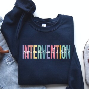 May include: A navy blue sweatshirt with the text "INTERVENTION specialist" in rainbow colors. The sweatshirt is laid out on a white surface with a pair of gray sneakers and blue jeans.