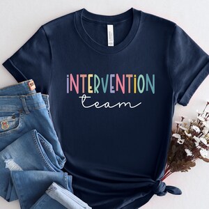 May include: A navy blue t-shirt with the text "intervention team" in rainbow colors. The shirt is folded and displayed on a white background with a pair of blue jeans and a bouquet of white flowers.