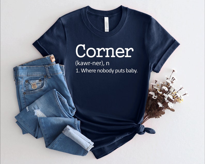 以下が含まれることがあります： 白い文字で「Corner (kawr-ner), n 1. Where nobody puts baby.」と書かれたネイビーブルーのTシャツです