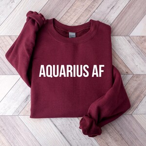 Op de afbeelding: Een bordeauxrood sweatshirt met de tekst "Aquarius AF" in wit gedrukt op de voorkant.