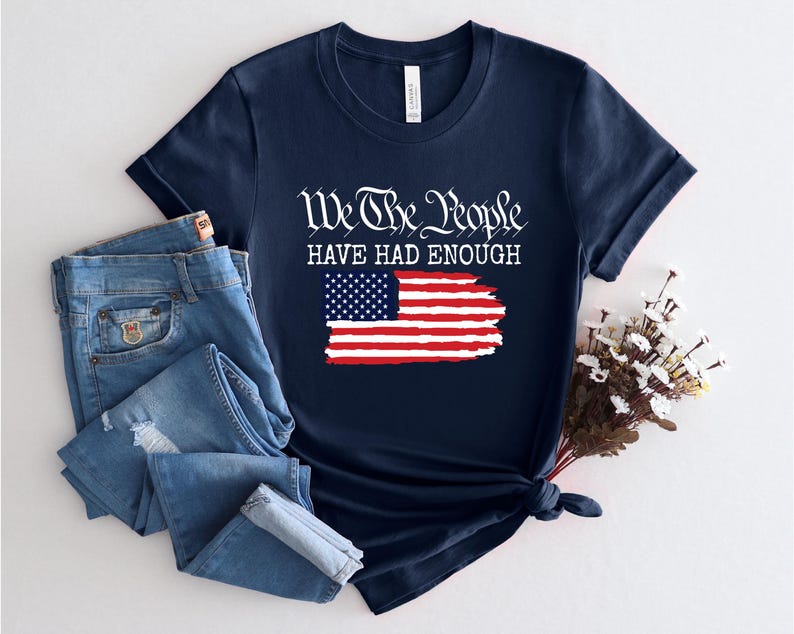 May include: A navy blue short-sleeve t-shirt featuring the white text We The People HAVE HAD ENOUGH above a graphic of the American flag with a distressed, brushstroke-style finish. The shirt is paired with blue denim jeans with distressed details.