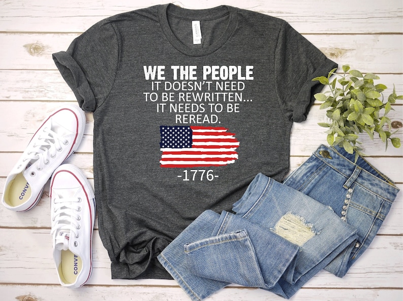 May include: A dark gray t-shirt with the text "WE THE PEOPLE IT DOESN'T NEED TO BE REWRITTEN... IT NEEDS TO BE REREAD. -1776-" and a distressed American flag graphic.