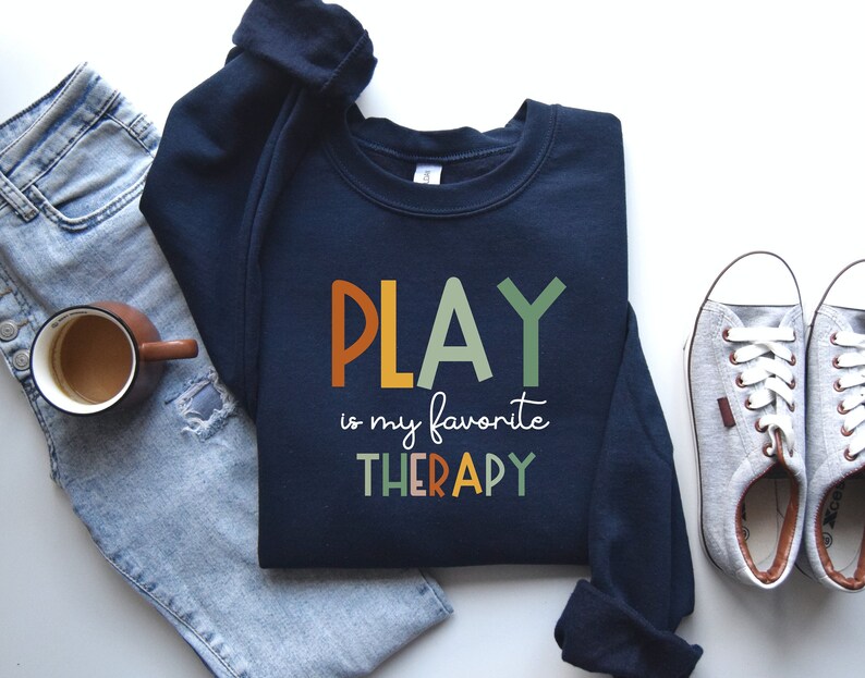 May include: A navy blue sweatshirt with the text "PLAY is my favorite THERAPY" in colorful letters. The letters are in a rainbow color scheme. The sweatshirt is laid flat on a white surface with a pair of jeans, a coffee mug, and a pair of white sneakers.