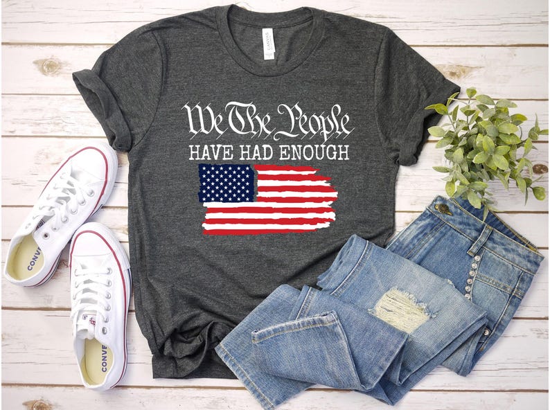 May include: Dark gray t-shirt with the text "We The People Have Had Enough" and a distressed American flag design. The image also includes white sneakers and blue jeans.