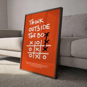 Może przedstawiać: Pomarańczowy plakat w ramie z białymi słowami "THINK OUTSIDE THE BOX" i grą w kółko i krzyżyk. Plakat opiera się o szarą sofę. Na dole plakatu znajduje się mały tekst.