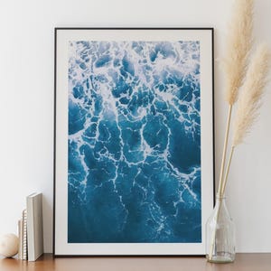Könnte beinhalten: Ein gerahmter Kunstdruck mit einer Luftaufnahme von Meereswellen. Das Bild zeigt eine tiefblaue Farbpalette mit weißen, schaumigen Mustern. Das Kunstwerk wird auf einem Holzschrank mit dekorativen Elementen wie einer Vase und Büchern ausgestellt.