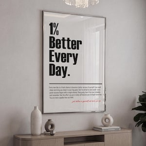 Peut inclure: A framed poster with the phrase "1% Better Every Day" in large, bold black letters. Below, smaller text offers motivational advice. The poster is displayed on a light wall above a wooden cabinet with decorative vases.