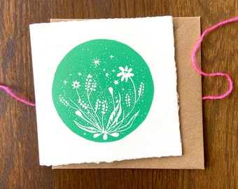 Könnte beinhalten: Eine weiße Karte mit einem grünen Kreis, der mit weißen Blumen und Sternen verziert ist. Die Karte hat einen rauen, strukturierten Rand.