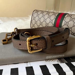 Könnte beinhalten: Ein brauner Leder-Handtaschenriemen mit goldfarbener Schnalle und Verschluss. Im Hintergrund eine passende Handtasche mit Markenlogo und grün-rotem Streifen.