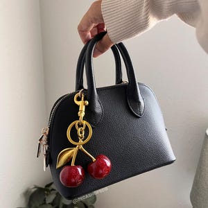 May include: A black, dome-shaped handbag with a gold-colored zipper and handles. A gold-colored cherry charm with red, glittery cherries and green leaves hangs from the handle. The bag has a textured surface.