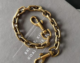 May include: A brass-colored chain with two clasp ends. The chain is made of rectangular links and is lying on a gray surface. The clasps are attached to the chain with a swivel mechanism. The text "BRI...GOAL...TE" is visible on the surface.