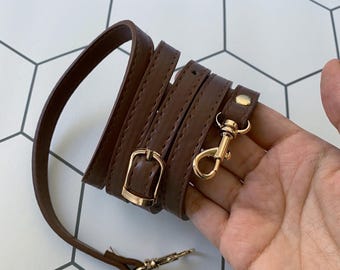 May include: A brown leather strap with gold-colored metal hardware, including a buckle and two swivel clasps. The strap is rolled up and appears to be new. The background is a white and gray geometric tile pattern.