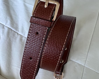 May include: A brown leather bag strap with a gold-colored buckle and clasp. The strap has a textured surface with white stitching along the edges. The buckle is rectangular and the clasp is a lobster claw style.