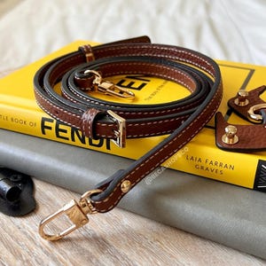 May include: A brown leather strap with gold hardware and stitching. The strap is coiled and laid on top of a yellow book with the word "FENDI" visible. The book is on top of a gray surface.