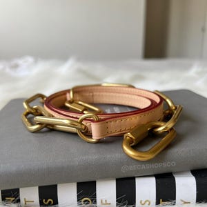 May include: A gold chain strap with a beige and red leather strap. The strap is laying on a gray book with black and white stripes.