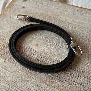 May include: A black leather purse strap with silver-colored metal clasps. The strap is coiled on a light-colored wooden surface. The strap is designed to be attached to a purse or bag.