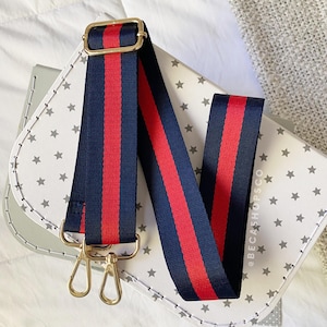 May include: A navy blue and red striped bag strap with gold-colored hardware. The strap is positioned over a white bag with a star pattern. The strap has a gold-colored buckle and two gold-colored clasps.