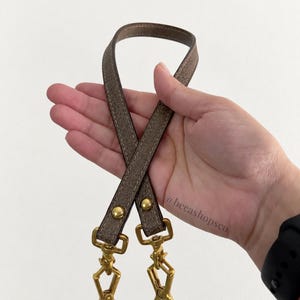 以下が含まれることがあります： 表面に質感のあるトープカラーのレザーストラップ。両端にはゴールドトーンの金属製回転式ロブスタークラスプが付いています。ストラップはゴールドトーンのリベットで固定されており、レザーの表面には「@becashopsco」という文字がプリントされています。
